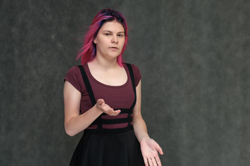 Portrait to the waist of a young pretty teenager girl in a burgundy T-shirt and dress with beautiful purple hair on a gray background in the studio. Talking, smiling, showing hands with emotions.