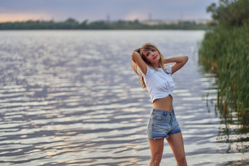 Fototapeta na wymiar slim girl in shorts and white T-shirt stands by the water on the lake