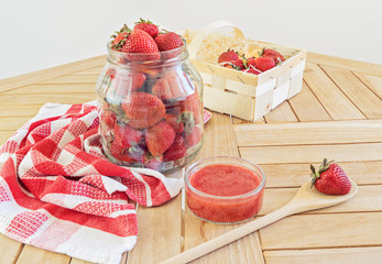 Large glass jar full of ripe strawberries and ready to eat strawberry jam with spoon on wooden...
