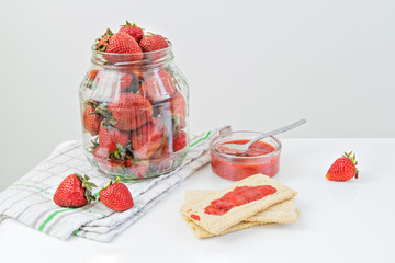 Large glass jar full of ripe strawberries and ready to eat strawberry jam on cracker with spoon....