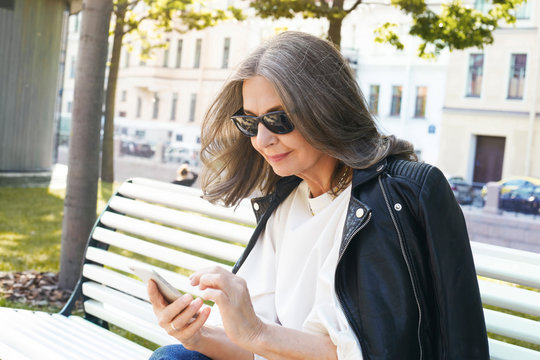 Outdoor picture of busy modern middle aged woman in stylish black sunglasses and leather jacket messaging online using smart phone, sitting on empty street in the morning. Technology and communication