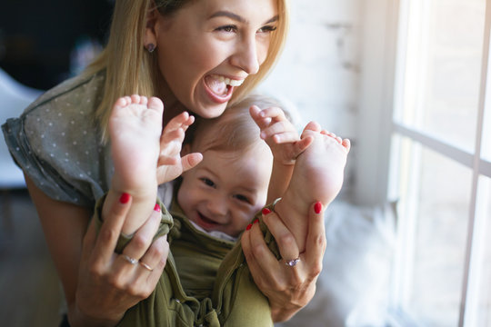 People, Family, Happy Childhood, Joy And Love Concept. Cheerful Attractive Young Woman Holding Feet Of Her Cute Baby Son, Opening Mouth Widely In Excitement, Having Fun Indoors. Posing By Window