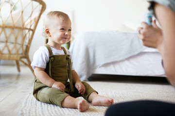 Picture of cute charming European infant sitting barefooted on carpet at home, having joyful excited facial expression, watching his mother blow soap bubbles. Childhood, joy, fun and happiness