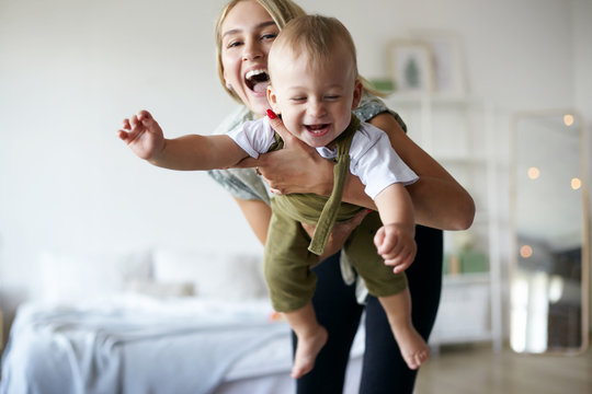 Pretty girl babysitting adorable toddler, having fun in stylish interior. Portrait of beautiful happy young Caucasian mom enjoying precious time with her cute baby son, holding him like flying plane