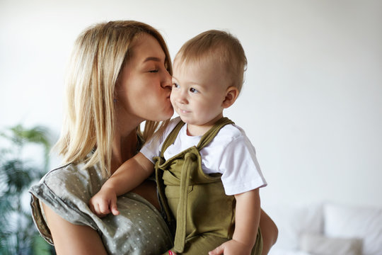 Indoor Shot Of Attractive Young European Mother With Blonde Hair Holding Tight Her Cute Chubby Ten Month Old Baby Son, Kissing Him On Cheek. Stylish Mom Bodning With Adorable Sweet Infant Child