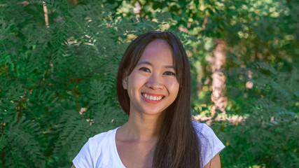 portrait asian woman with friendly smile in the park. Happy young girl standing in front of green tree outdoors. Vietnamese female wearing in casual t-shirt smiling looking at the camera. Her long