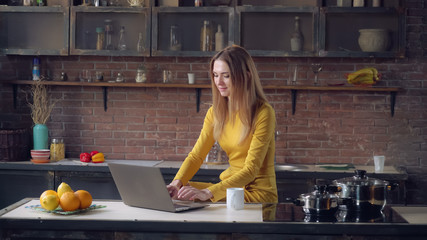 laughing businesswoman working at home. Young female wearing in elegant dress sitting at cooking table working on laptop.