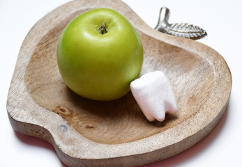 An image of a green apple and a model of a human molar of a white color on a wooden plate in the apple form on a white surface