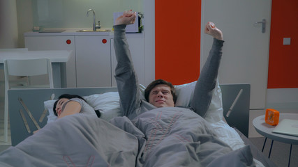 husband awake in the morning holding alarm clock next to lying his wife in bed with white pillows....
