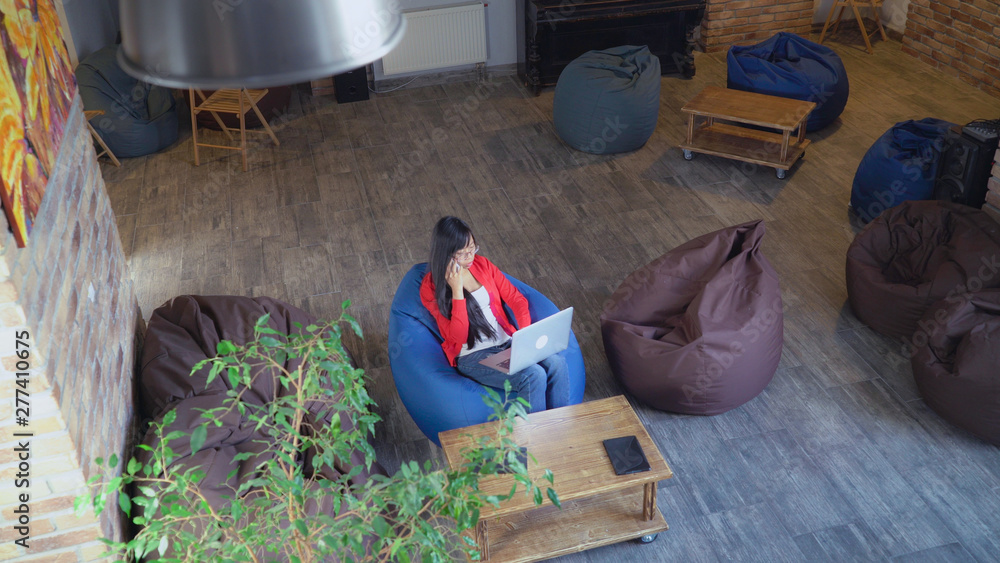 Poster Emptyin startup company only one woman sitting in casual office calling client and checking information in computer. Asian young female wearing in stylish red cardigan has phone conversation use