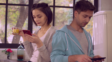 Two people using gadgets in the morning on the kitchen. behind young happy family big window with autumn view. Couple in love talking, smiling, surfing internet or reading news or sharing in social