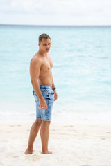 A strong young guy with a bare-chested walks on a white sandy beach on a tropical island