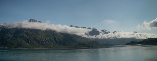 wispy clouds onto of snowy mountain peaks, with reflection in teal glacier waters in Alaska. 
