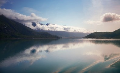 Fototapeta na wymiar wispy clouds onto of snowy mountain peaks, with reflection in teal glacier waters in Alaska. 