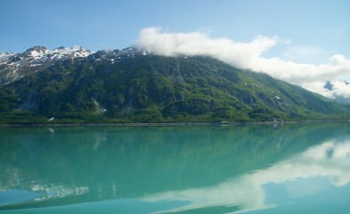 Naklejka na ściany i meble wispy clouds onto of snowy mountain peaks, with reflection in teal glacier waters in Alaska. 