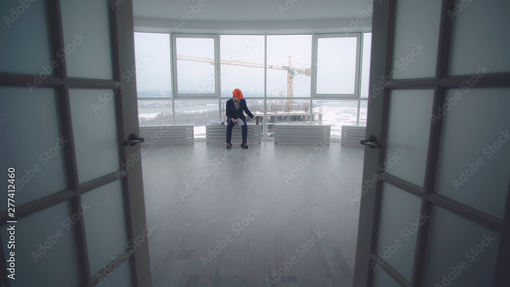 Poster tired handsome architect has a break resting drinking coffee. Manager of building engineer sitting in the modern office real estate company. Handsome man wearing in elegant suit and orange hard hat.