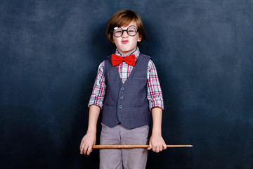 Funny intelligent smart schoolboy boy with eye glasses holding wooden teacher pointer opposite school chalkboard at school. Back to school, ready to study concept