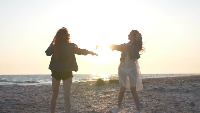 sunset rays over the sea illuminate the dance on the beach of two young sexy women dressed in boho style