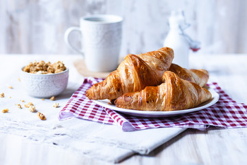 Delicious breakfast french style at home: light background, white plate with fresh croissants, small bowl of granola, cup of tea and bottle with milk. Served on red and beige napkins