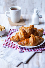 Delicious breakfast french style at home: light background, white plate with fresh croissants, small bowl of granola, cup of tea and bottle with milk. Served on red and beige napkins