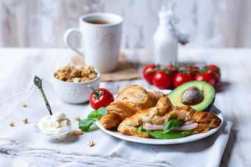 Delicious breakfast french style at home: light background, white plate with fresh croissants, small bowls of granola and cream cheese, cup of tea, avocado, tomatoes, basil. Served on beige napkins