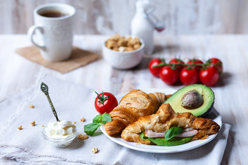 Delicious breakfast french style at home: light background, white plate with fresh croissants, small bowls of granola and cream cheese, cup of tea, avocado, tomatoes, basil. Served on beige napkins
