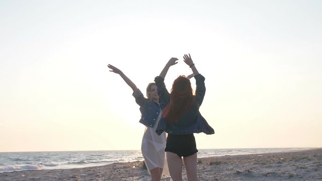 two young sexy women dressed in the style of boho rejoice and have fun in the rays of the sunset over the sea on the beach