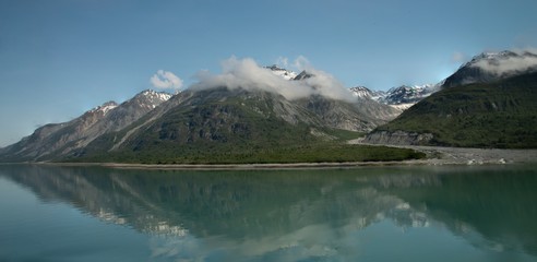 Obraz na płótnie Canvas wispy clouds onto of snowy mountain peaks, with reflection in teal glacier waters in Alaska. 