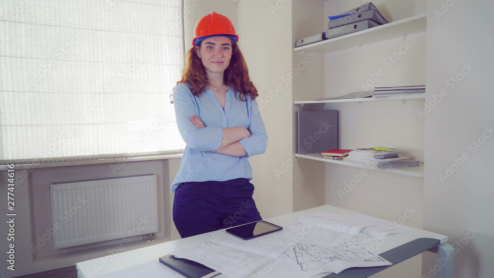 Poster Redheaded engineer at work using digital tablet. Professional female looking at the camera with arms crossed. On the desk sketch with technical drawing some building.