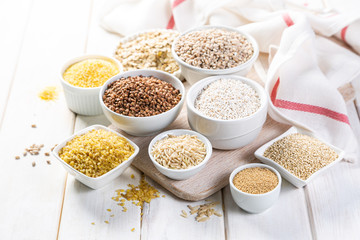 Selection of whole grains in white bowls - rice, oats, buckwheat, bulgur, porridge, barley, quinoa, amaranth, on white wood background