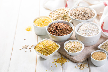 Selection of whole grains in white bowls - rice, oats, buckwheat, bulgur, porridge, barley, quinoa,...