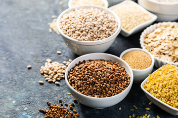 Selection of whole grains in white bowls - rice, oats, buckwheat, bulgur, porridge, barley, quinoa, amaranth on dark background