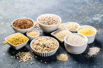 Selection of whole grains in white bowls - rice, oats, buckwheat, bulgur, porridge, barley, quinoa, amaranth on dark background