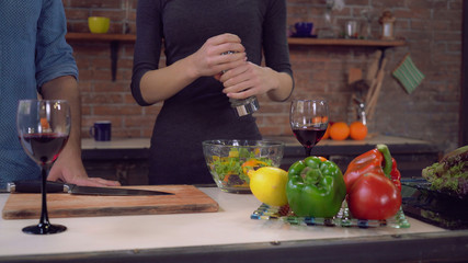 Woman cooking in the kitchen. Female hand holding handheld pepper mills. Lady peppering salad in a bowl. Man taking wineglass with red wine. On the cook table different vegetables and fruits. People
