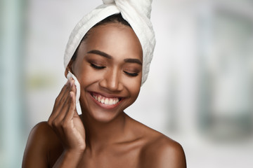 Close-up portrait of a beautiful smiling woman with clean skin cleaning her face with cotton swab pad isolated on bright background. Beauty treatments and cosmetics spa therapy