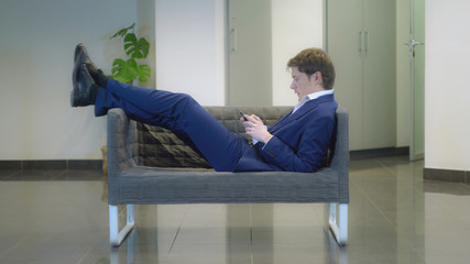 Portrait man wearing in fashionable elegant blue suit resting on the sofa playing game or texting message on smartphone. Caucasian businessman using wireless in casual lobby.