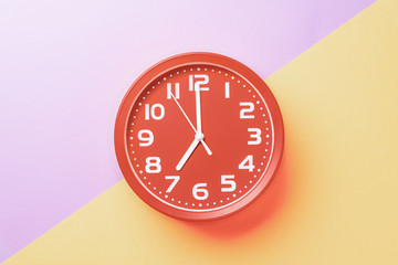 Red clock showing seven o'clock with white numbers on a yellow lilac background