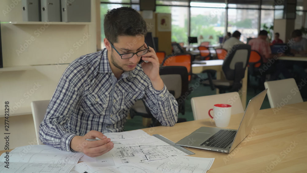 Poster engineer sitting in at the working place on the desk blueprint. young professional man call client t