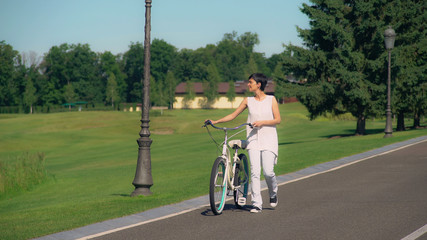 Young brunette resting outdoors at summertime. Happy smiling woman going with bike looking on nature view with green grass and trees. Cheerful girl with short hair in city with bicycle.