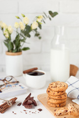 Morning breakfast. A cup of coffee with cinnamon, tasty cookies with chocolate, a bottle of milk, a bouquet of flowers and a book with glasses on a white brick wall background