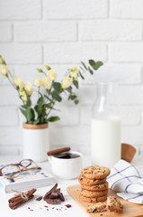 Morning breakfast. A cup of coffee with cinnamon, tasty cookies with chocolate, a bottle of milk, a bouquet of flowers and a book with glasses on a white brick wall background with space for text