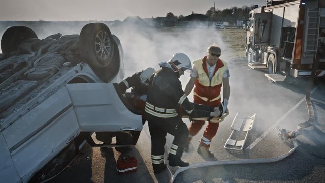 On the Car Crash Traffic Accident Scene: Rescue Team of Firefighters Pull Female Victim out of Rollover Vehicle, They Use Stretchers Carefully, Hand Her Over to Paramedics who Perform First Aid