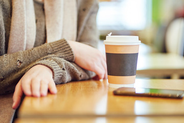 woman in a cafe with a paper cup of coffee. brown sweater and scarf. sits by the table. close up.