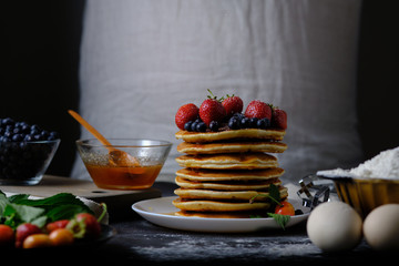 Pancakes decorated with blueberries and strawberries. On the table are the products from which they prepared the dish and berries. Dad prepares a delicious breakfast. Sugar powder in flight.