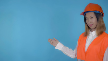 young asian woman posing pointing on copy space on blue background in studio . attractive millennial girl wearing white casual shirt and orange hard hat and vest looking at the camera with cheerful