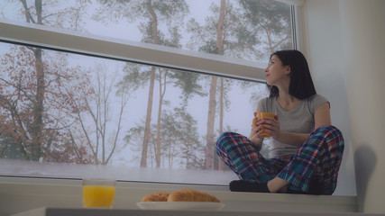 Portrait young woman enjoy loneliness in hotel room or at home in winter season. Attractive brunette drinking coffee and looking through the window on beautiful snowing view.