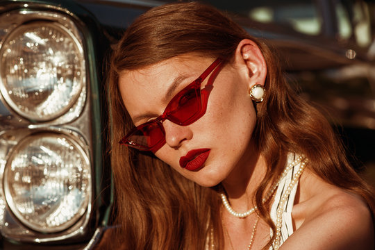 Outdoor close up portrait of young beautiful woman with freckles, red lips, wearing red color trendy sunglasses, pearl earrings, necklace, model posing near retro car