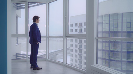 businessman in empty room with big panoramic windows looking on the urban city view. young professional man wearing in elegant suit alone in modern apartment. caucasian male deep in thought.