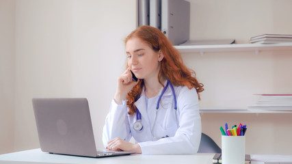 Doctor working with medical records on computer talking by phone. Professional redhead therapist wearing in lab white coat and phonendoscope check information or surfing internet has conversation with