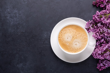 Cup of morning coffee with spring lilac flowers branches blossoming on black stone background Top...